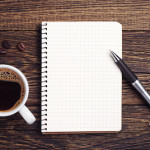 A cup of black coffee, a notepad, and a pen on a wooden table