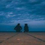 Two people wearing hoodies sitting on the floor with a candle between them and a beautiful blue cloudy sky above them
