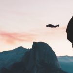 Man jumping off a cliff with mountains and colourful sky in the background